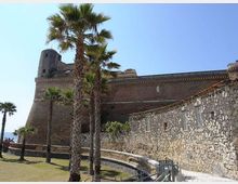 Fortezza costiera in mattoni con bastioni e torre angolare, affiancata da un lungo edificio in pietra con piccole finestre. In primo piano ci sono palme e un prato con percorso pedonale e ringhiera, sotto un cielo sereno.