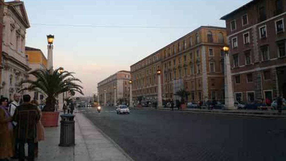 Ampio viale urbano al tramonto con carreggiata in asfalto scuro e marciapiede in pietra, fiancheggiato da lampioni e grandi palme in vaso. Ai lati si trovano alti edifici storici in muratura con facciate uniformi e finestre regolari, con alcune auto in transito lungo la strada.