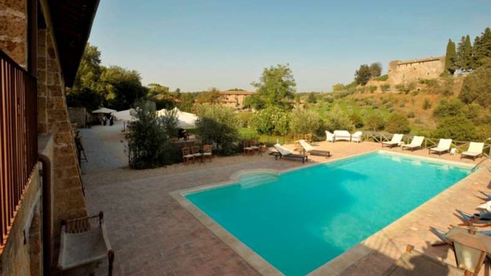 Piscina rettangolare all’aperto con bordo in pietra e pavimentazione in cotto, circondata da lettini e ombrelloni su una terrazza. Sullo sfondo si vede un paesaggio collinare con alberi e un edificio in pietra, accanto a una struttura rustica con balcone.