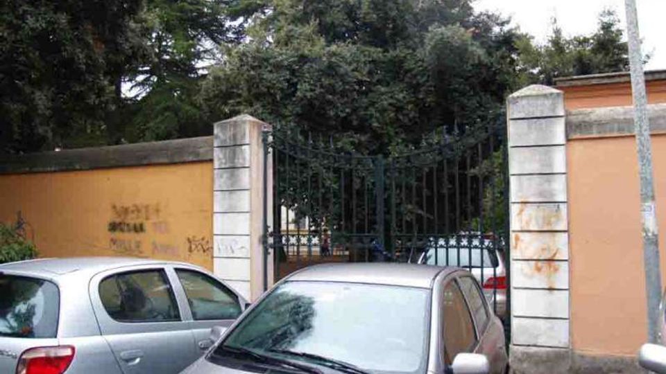 Ingresso con cancello in ferro battuto tra pilastri in pietra e muro intonacato color ocra, affacciato su una strada con auto parcheggiate; dietro il cancello si vede un’area verde con alberi alti e fitti.