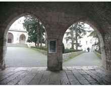 Passaggio coperto in pietra con due grandi archi che si affacciano su un cortile lastricato e vialetti in ciottoli. Sullo sfondo si vedono edifici storici chiari con porticati e un ingresso ad arco, affiancati da alberi e muretti in pietra.