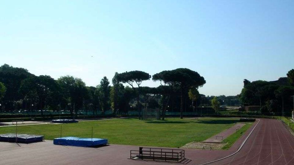 Pista di atletica all’aperto con corsie rosse che curva attorno a un grande prato centrale, con pedane e materassi per salto in alto. Sullo sfondo si vedono alberi alti e un cielo sereno.