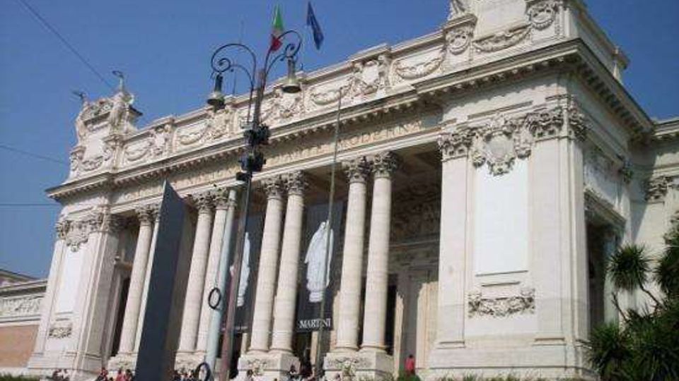 Facciata di un grande edificio monumentale neoclassico con alto portico e colonne corinzie, ricchi bassorilievi e timpano decorato. Davanti si vedono una scalinata, lampioni stradali e bandiere sul tetto, in un contesto urbano.