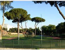 Vista di un impianto sportivo all’aperto con pista di atletica rossa che circonda un campo in erba, recintato da una rete metallica. Sullo sfondo si vedono alti pini a ombrello e altri alberi, con alcuni edifici e pali dell’illuminazione ai margini.