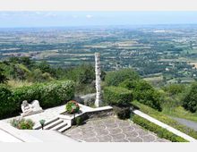 Terrazza panoramica su una collina con pavimentazione decorata, gradini e aiuole fiorite, affacciata su una vasta pianura con campi e aree boschive. Al centro si vede un alto monumento in pietra e una scultura bianca vicino al parapetto.