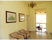 Interno di un corridoio o salottino con pareti chiare, due poltrone imbottite e quadri incorniciati appesi; dal soffitto pende un lampadario classico. In fondo, una porta-finestra aperta conduce a una veranda luminosa con sedie e vista sull’esterno.