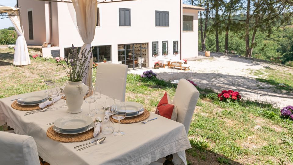 Tavolo da pranzo all’aperto apparecchiato sotto un gazebo con tende bianche, in un giardino con aiuole fiorite. Sullo sfondo si vede una casa di campagna chiara con finestre scure e area esterna pavimentata, circondata da alberi.