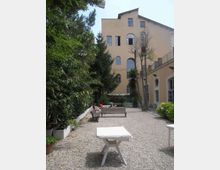 Cortile interno con pavimentazione in ghiaia, panchine e tavoli bianchi, circondato da alberi e siepi. Sullo sfondo si vede un edificio chiaro a più piani con grandi finestre ad arco e, a destra, una struttura con finestre e scala esterna.