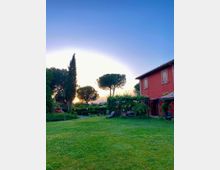Ampio giardino con prato verde e alberi alti, con lampioni e sedute lungo un vialetto. Sulla destra si vede una casa di campagna a due piani con facciata rossa, persiane in legno e portico coperto da rampicanti.