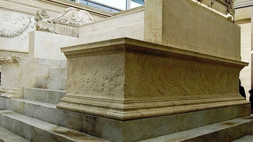 Interno di museo luminoso con grandi finestre e soffitto a lucernario, dove un grande basamento a gradoni in pietra sostiene un sarcofago monumentale scolpito con motivi ornamentali e figure. Pareti in pietra chiara con rilievi decorativi lungo i lati.