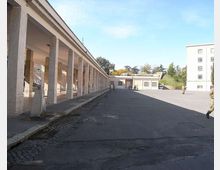Ampio cortile asfaltato con un lungo porticato a sinistra, caratterizzato da una fila di colonne in muratura e soffitto piatto. Sullo sfondo si vedono edifici bassi e un palazzo a più piani, con alberi oltre il complesso sotto un cielo sereno.
