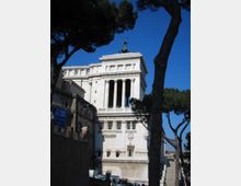 Edificio monumentale in pietra chiara con facciata neoclassica, grandi colonne e loggiato, sormontato da una statua sul tetto. La struttura è vista dal basso ed è incorniciata da alti pini contro un cielo azzurro.