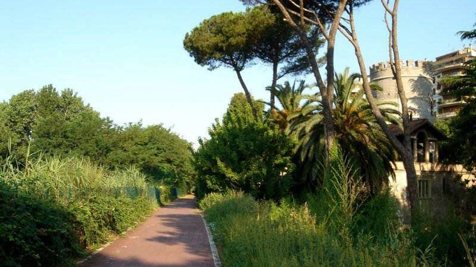 Sentiero asfaltato in un’area verde, circondato da vegetazione alta e alberi mediterranei come pini e palme. Sullo sfondo si intravede un edificio in pietra con torretta circolare e merli, parzialmente nascosto tra gli alberi.