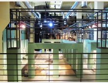 Interno di una sala espositiva in stile industriale, con travi e tubature a vista sul soffitto e una struttura metallica sospesa. Pareti verde acqua delimitano aree con reperti e pavimenti decorati, osservabili da una balconata con ringhiera in primo piano.
