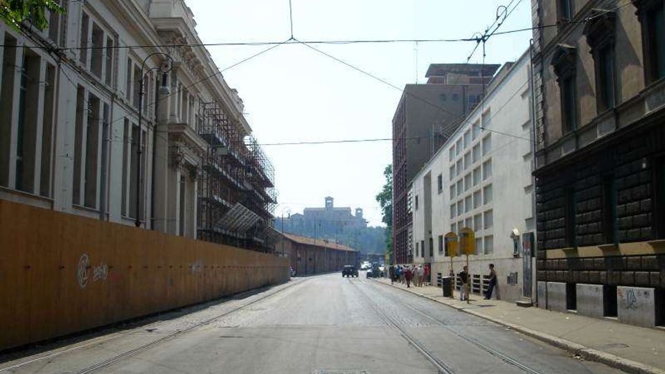 Strada urbana ampia tra edifici alti, con binari del tram sulla carreggiata e cavi aerei sospesi. Ai lati si vedono facciate storiche e moderne, un lungo cantiere recintato e marciapiedi con segnaletica e lampioni.