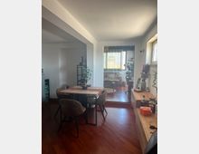 Interno di un appartamento con zona pranzo open space, tavolo rettangolare con quattro sedie su pavimento in legno scuro e pareti bianche. Sullo sfondo, un piccolo salotto rialzato con divano e grande finestra con tende, affiancato da piante e mobili bassi lungo la parete.