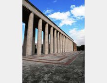Lungo colonnato monumentale in pietra con alte colonne cilindriche che sorreggono un’ampia trabeazione, affacciato su una grande piazza pavimentata con motivi geometrici. Sullo sfondo si vede un cielo azzurro con nuvole e una fascia di alberi ai margini.