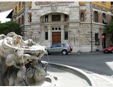 Fontana in pietra con sculture e getti d’acqua in una vasca circolare, in primo piano su una strada urbana. Sullo sfondo si vede la facciata di un edificio storico con portale in legno, decorazioni in pietra e finestre ad arco.