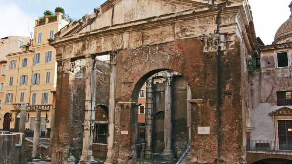 Rovine di una struttura monumentale in mattoni e pietra con grande arco centrale e colonne parzialmente conservate, affacciata su una piazza urbana. Sullo sfondo si vedono edifici residenziali con facciate chiare e una cupola.