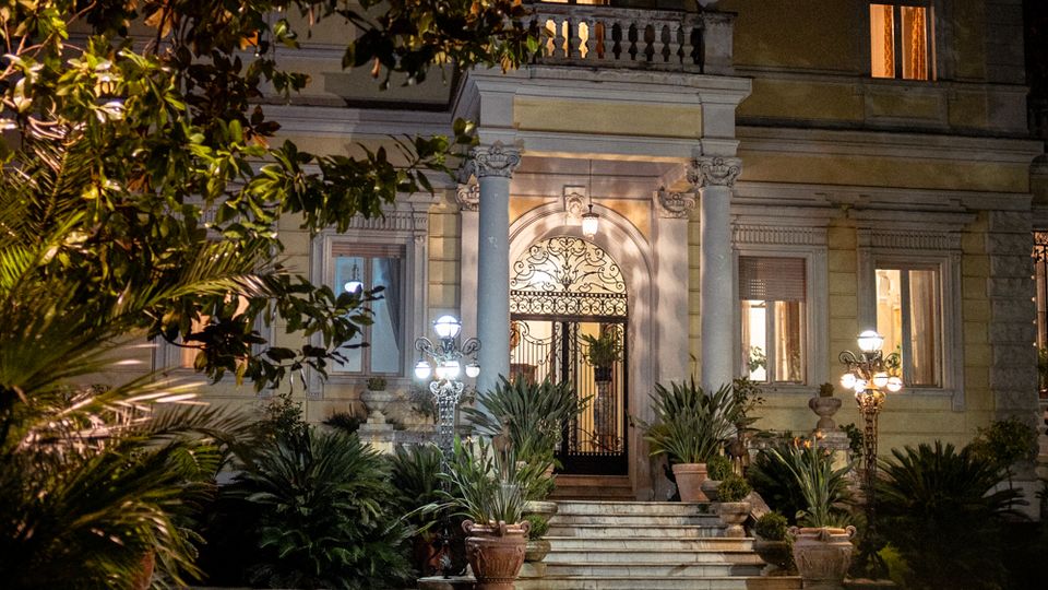 Facciata notturna di una villa storica con ingresso ad arco, cancellata in ferro battuto e scalinate illuminate da lampioni. Davanti si estende un giardino con piante ornamentali e vasi ai lati dei gradini.