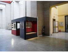 Ampio atrio interno con pavimento in pietra e alte pareti in cemento chiaro illuminate da lucernari. Al centro è presente un chiosco espositivo moderno nero e rosso con vetrine e pannelli, accanto a un passaggio verso un corridoio giallo con porte e grate.