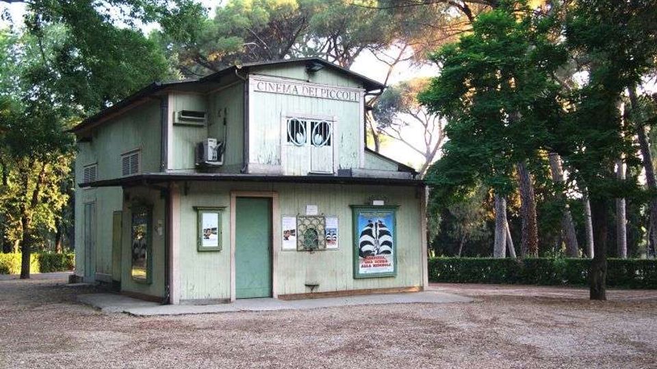 Piccolo edificio in legno a due piani, con facciata chiara e tetto sporgente, con insegna “CINEMA DEI PICCOLI” e locandine alle pareti. La struttura è immersa in un parco con alti alberi e un ampio spazio sterrato antistante.