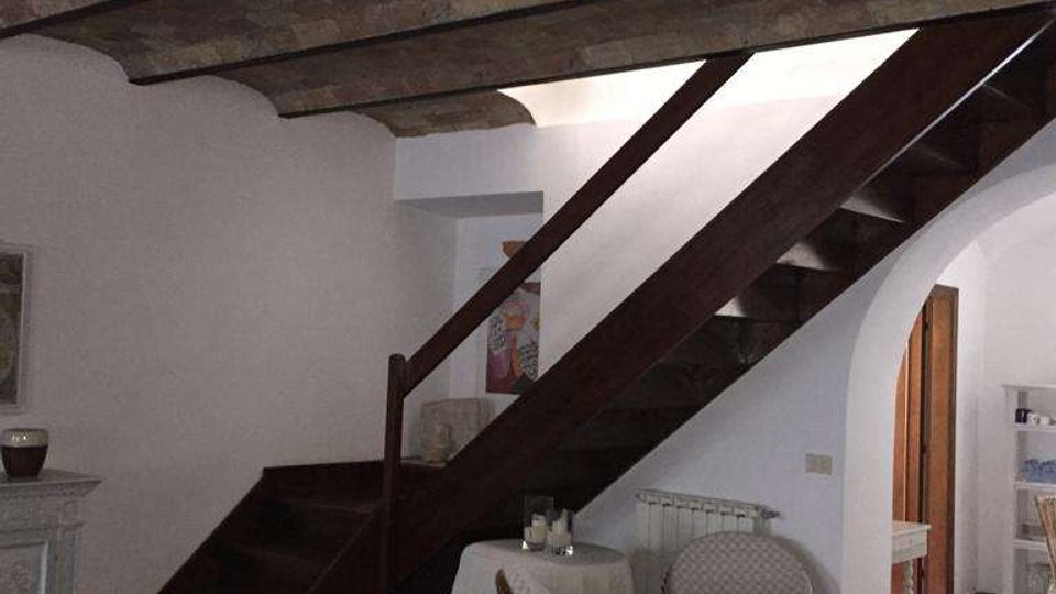 Interno di una casa con soffitto a travi e voltine in mattoni e pavimento in cotto, dominato da una scala in legno scuro che sale lungo la parete. Sotto la scala c’è un piccolo angolo con poltrone in vimini e tavolino rotondo, accanto a un arco che conduce a un’altra stanza.
