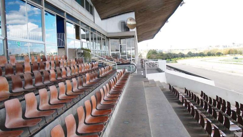 Tribuna di un impianto sportivo con lunghe file di sedili in plastica marrone su gradoni in cemento, affiancata da un edificio con ampie vetrate. Un grande tetto a sbalzo copre le gradinate e, sul lato destro, si intravede la pista o area di gara all’aperto.