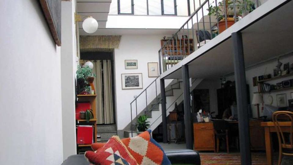 Interno di un loft su due livelli con soppalco e ringhiera metallica, illuminato da una grande vetrata a soffitto inclinata. Al piano terra si vedono divano e area studio, mentre una scala conduce al livello superiore con piante e arredi.