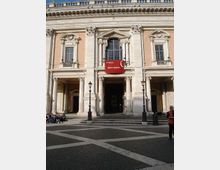 Facciata di un edificio storico in stile classico con portico a colonne e tre ingressi ad arco, grandi finestre incorniciate e decorazioni in pietra chiara su intonaco rosato. Davanti si vede una piazza lastricata con lampioni, e al centro un drappo rosso con la scritta “Musei Capitolini” appeso sotto la finestra.