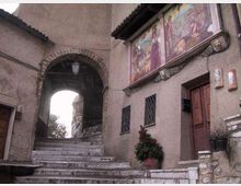 Scalinata in pietra che conduce a un arco in muratura con lanternino, tra edifici intonacati. Sulla facciata a destra sono visibili grandi affreschi sotto il tetto, finestre con inferriate e una porta in legno.