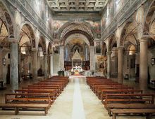 Interno di una chiesa a tre navate con file di banchi in legno lungo una navata centrale che conduce all’altare. Colonne in pietra e arcate laterali sostengono volte decorate, con affreschi sulle pareti e soffitto a cassettoni.