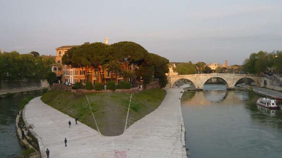 Veduta di un’isola fluviale con una passeggiata in pietra che segue la riva, alberi fitti e edifici storici. Sullo sfondo si vede un ponte in muratura a più arcate che attraversa il fiume.