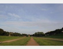 Ampio prato in un parco con un sentiero sterrato centrale che conduce verso edifici lontani all’orizzonte. Ai lati si vedono alberi e lievi pendii erbosi sotto un grande cielo azzurro con nuvole sottili.
