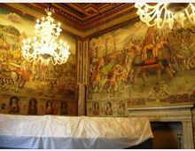 Salone interno con pareti affrescate raffiguranti scene storiche e figure, soffitto a cassettoni decorato e grandi lampadari in vetro sospesi. In basso si vede un ampio telo bianco che copre parte della sala e una grande cornice architettonica simile a un camino.