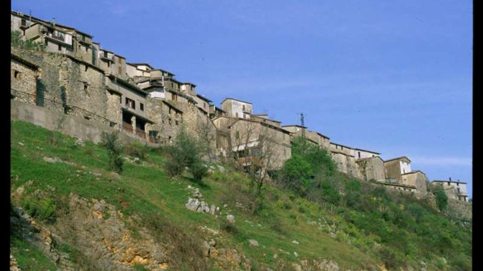 Case in pietra di un borgo arroccato lungo un pendio erboso, con edifici addossati tra loro e muri in muratura a vista. In primo piano si vede il versante roccioso con cespugli e alberi sparsi sotto un cielo sereno.