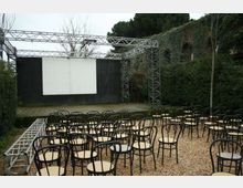 Piccolo teatro all’aperto con palco e grande schermo bianco incorniciato da pannelli neri e struttura metallica a traliccio. Davanti al palco ci sono file di sedie in metallo su ghiaia, circondate da siepi e vegetazione.