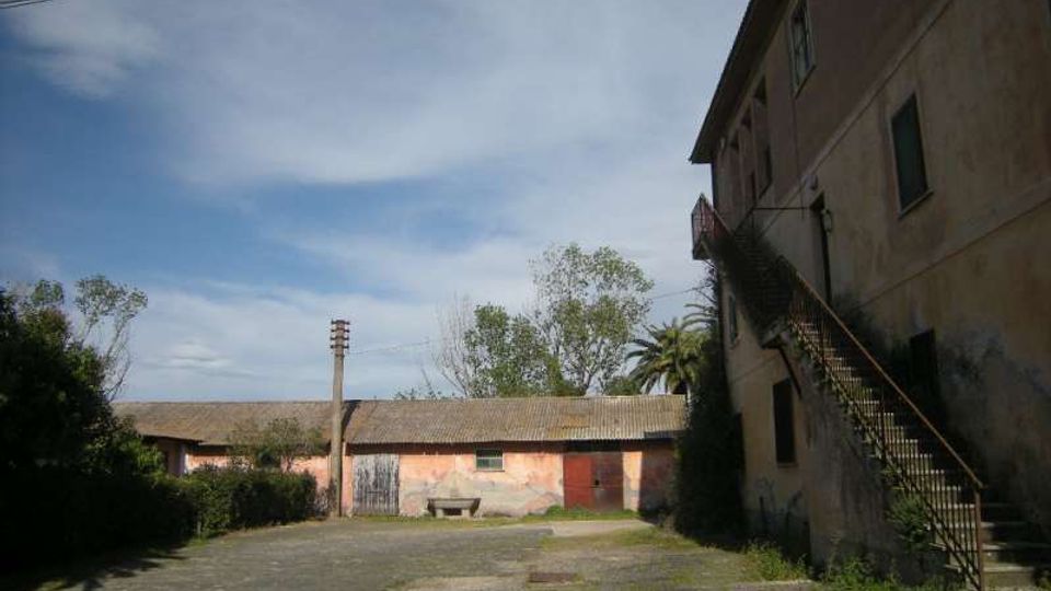 Cortile esterno in cemento davanti a edifici rurali, con un basso fabbricato intonacato e tetto a falda sullo sfondo e un edificio più alto a destra con scala metallica esterna. Sullo sfondo si vedono alberi e un cielo parzialmente nuvoloso, con cavi elettrici sospesi.