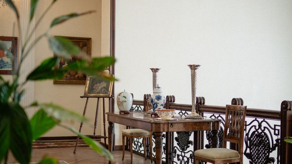 Interno di un salone in stile classico con pavimento in parquet, soffitto decorato in stucco e lampadario; lungo una ringhiera in ferro battuto ci sono un tavolo con ceramiche e candelieri e sedie imbottite. Sullo sfondo si vede un passaggio verso un’altra stanza con quadri e cavalletto, con una pianta in primo piano sfocata.