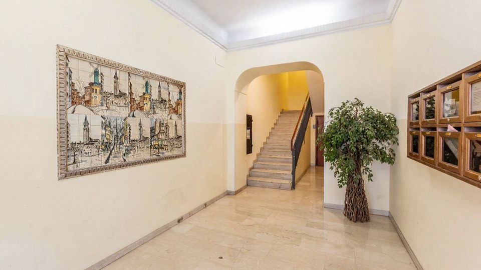 Ingresso condominiale con pavimento in marmo chiaro e pareti color crema, con un grande quadro decorativo sulla sinistra e una fila di cassette postali in legno sulla destra. In fondo, un arco conduce a una scala in pietra con corrimano scuro; accanto è presente una grande pianta d’interno.