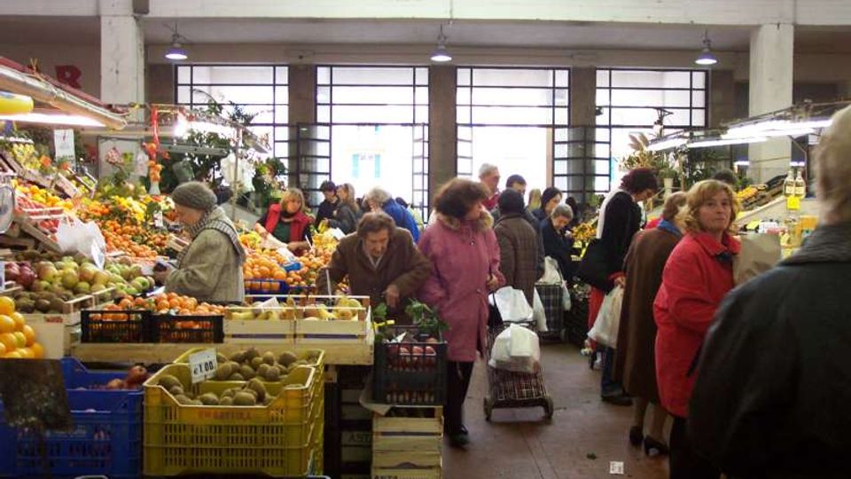 Interno di un mercato coperto con banchi di frutta e verdura su entrambi i lati, casse e cassette impilate lungo il corridoio centrale. Sul fondo si vedono grandi finestre a griglia e illuminazione a sospensione sotto un soffitto alto.