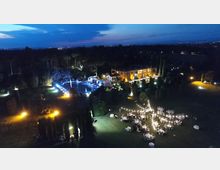 Veduta aerea notturna di una villa con giardini alberati, piscina illuminata e aree esterne con tavoli e luci decorative. Sullo sfondo si estende un paesaggio boscoso con luci urbane all’orizzonte sotto un cielo al crepuscolo.