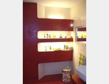 Bagno moderno con parete bordeaux e nicchie orizzontali illuminate usate come mensole per prodotti. A destra si vedono uno specchio con luce a braccio, un lavabo bianco e un piccolo sgabello.