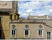 Facciata di un edificio storico intonacato con tre finestre ornate da cornici in stucco e motivi decorativi sopra gli architravi, sotto un parapetto del tetto. Sullo sfondo si vedono tetti in tegole e, in lontananza, una torre cittadina sotto un cielo parzialmente nuvoloso.