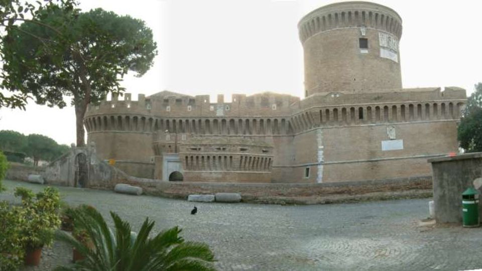 Veduta esterna di una grande fortificazione in mattoni con mura merlate e un’alta torre cilindrica, affacciata su una piazza lastricata. Intorno si vedono alberi ad alto fusto e vegetazione, con un basso muro di contenimento lungo il perimetro.