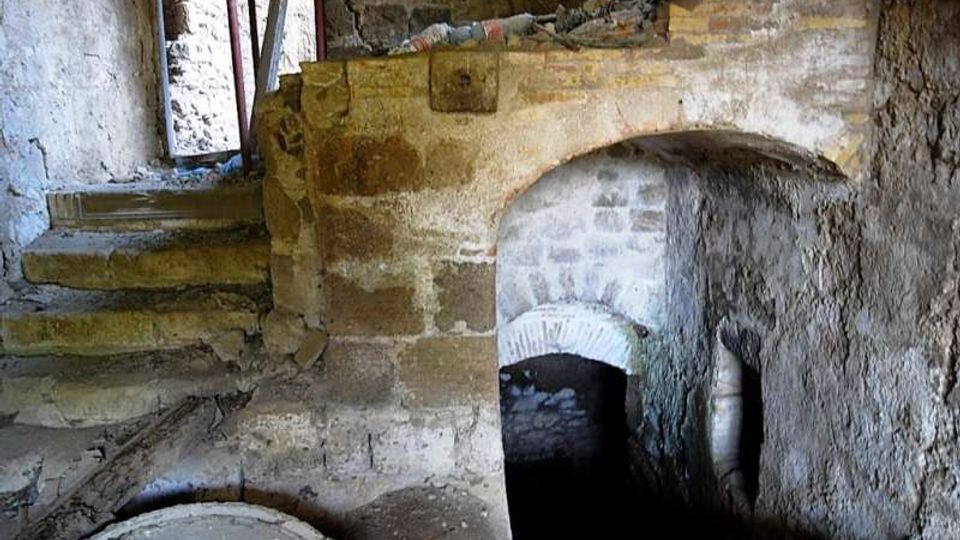 Interno di un ambiente sotterraneo in pietra e mattoni, con arco ribassato che conduce a un passaggio buio e una breve scala laterale in muratura. Si vedono pareti grezze, detriti sul pavimento e una grande apertura circolare in primo piano.