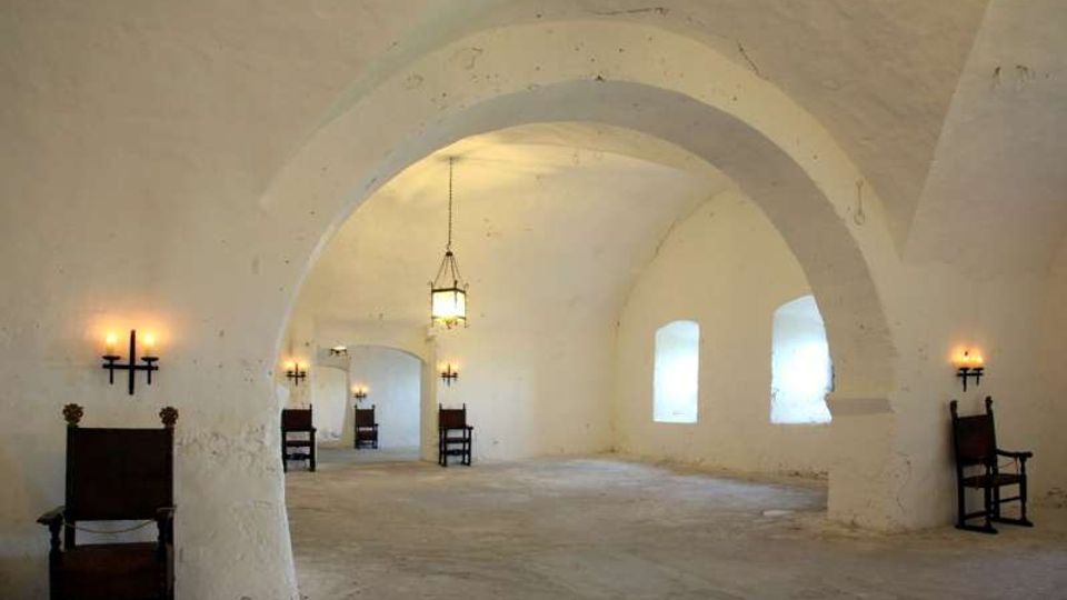 Ampia sala con soffitto a volte bianche e grandi archi in muratura, pavimento in pietra e finestre rettangolari lungo la parete destra. L’ambiente è illuminato da un lampadario centrale e da applique a parete, con alcune sedie in legno disposte lungo i lati.