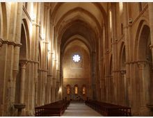 Interno di una chiesa in pietra con navata centrale lunga, alte arcate e colonne massicce che sostengono volte a crociera. In fondo si vede una grande finestra a rosone sopra tre finestre più piccole e file di panche ai lati del corridoio.