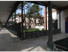 Portico lungo con pavimento piastrellato e tettoia sorretta da colonne metalliche, affiancato da una recinzione bassa. Sullo sfondo si vede un cortile con alberi alti e un edificio in muratura e intonaco con archi e dettagli in mattoni.