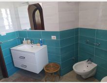 Bagno moderno con pareti piastrellate bianche e fascia turchese, lavabo con mobile sospeso e grande specchio. A destra si vede un bidet e una barra portasciugamani, con piccolo sgabello in vimini sul pavimento.